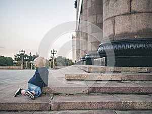 A little girl crawls on her knees along the granite steps of a huge building. Monumental granite columns rise in front