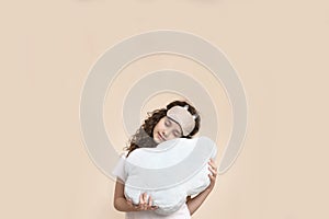 Little girl with curly hair isolated on a beige background, holding a pillow, have a sleeping mask.