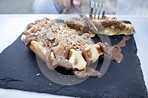 Little girl cuts waffle covered with dark chocolate topping and cereals powdered