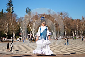 a little girl dancing flamenco dressed in a white dress with ruffles and blue fringes in a famous square in seville, spain. The photo