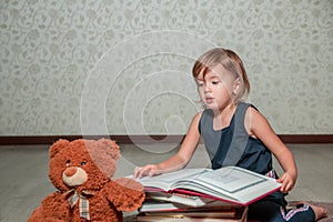 little girl in dark blue dress reading book sitting on the floor near teddy bear. Child reads story for toy.