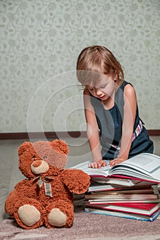 little girl in dark blue dress reading book sitting on the floor near teddy bear. Child reads story for toy.