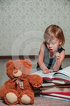 little girl in dark blue dress reading book sitting on the floor near teddy bear. Child reads story for toy.