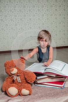 Little girl in dark blue dress reading book sitting on the floor near teddy bear. Child reads story for toy.