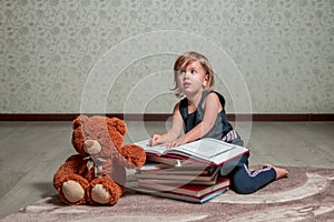 Little girl in dark blue dress reading book sitting on the floor near teddy bear. Child reads story for toy.