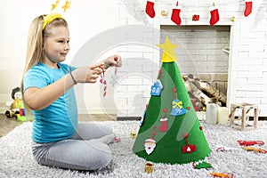 A little girl decorates a felt tree with New Year`s toys in a room near a fireplace