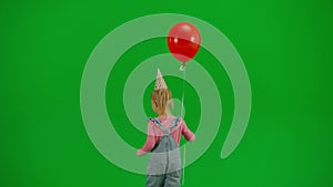 A little girl in denim overalls with a festive cap on her head and carrying a red helium balloon walks in front of a