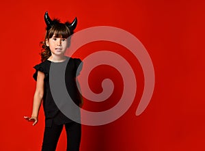 Little girl with devil horns, in black blouse and leggings posing against red studio background. Masquerade, Halloween. Close up