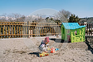 Little girl digs sand on the playground in the park near the toy house