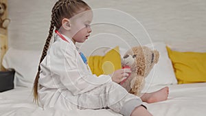 little girl doctor in white coat treats sick teddy bear, child veterinarian listens with stethoscope to patient, kid