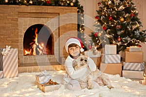 Little girl and dog at Christmas Eve sitting on white soft carpet, child looking at camera and hugging her pet, female kid in