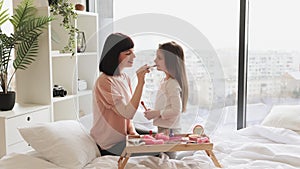 Little girl doing makeup to mom sitting at bedroom.