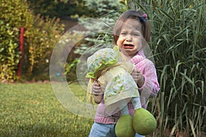 A little girl with a doll in her hand, standing next to tall grass and crying.