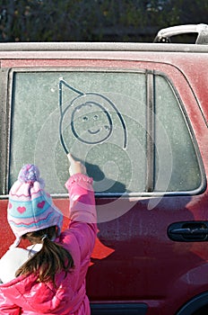 Little girl draw a happy face on a frosted car window