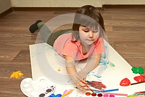 A little girl draws a watercolor on a white sheet of paper. She`s lying on the floor. Copy  space