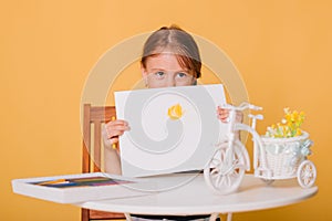 Little girl draws on a white sheet of paper. A creative child shows the sun hatch. Studio photo on a yellow background. art