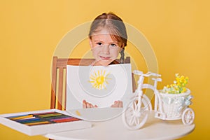 Little girl draws on a white sheet of paper. A creative child shows the sun hatch. Studio photo on a yellow background. art