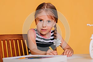 Little girl draws on a white table with crayons. Creative child. Studio photo on a yellow background. Art