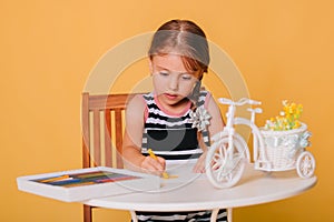 Little girl draws on a white table with crayons. Creative child. Studio photo on a yellow background. Art