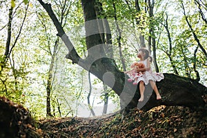 Little girl in a dress with doll sits on a tree in the forest