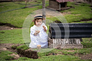 little girl in a dress and hat sits near a bird house on a farm holding an egg in her hand