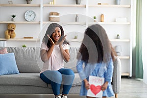 Little girl in dress hiding greeting card for mother's day behind her back, excited mom happy over surprise at home