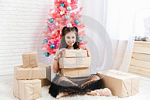 Little girl in a dress sits on the floor at home near the Christmas pink tree and holds boxes with gifts