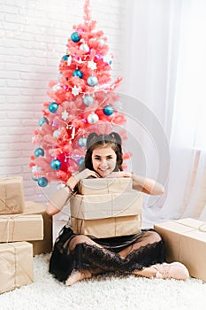 Little girl in a dress sits on the floor at home near the Christmas pink tree and holds boxes with gifts