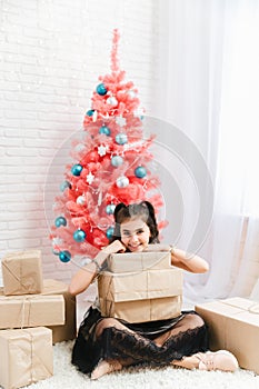 Little girl in a dress sits on the floor at home near the Christmas pink tree and holds boxes with gifts
