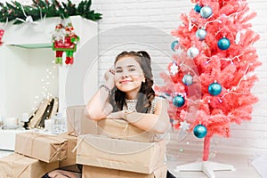 Little girl in a dress sits on the floor at home near the Christmas pink tree and holds boxes with gifts