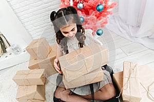 Little girl in a dress sits on the floor at home near the Christmas pink tree and holds boxes with gifts