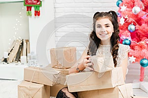 Little girl in a dress sits on the floor at home near the Christmas pink tree and holds boxes with gifts