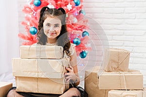 Little girl in a dress sits on the floor at home near the Christmas pink tree and holds boxes with gifts