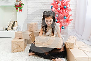 Little girl in a dress sits on the floor at home near the Christmas pink tree and holds boxes with gifts