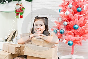 Little girl in a dress sits on the floor at home near the Christmas pink tree and holds boxes with gifts