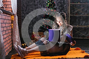 Little girl in a dress sits on the floor at home near the Christmas tree and holds a box with gifts