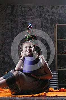 Little girl in a dress sits on the floor at home near the Christmas tree and holds a box with gifts