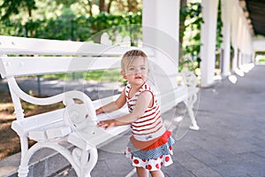 Little girl in a dress stands near a white bench in a long pavilion in the park