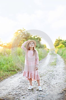 Little girl in dress and straw hat is walking along country road. Happy childhood. Summer vacation. Kid walks along rural road on