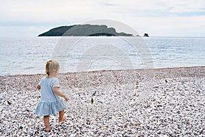 Little girl in a dress walks along a pebble beach to the sea with an island in the distance
