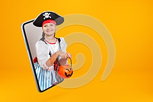 A little girl dressed as a pirate for Halloween holds a bucket with a pumpkin lantern and looks from a smartphone.