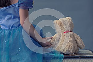 A little girl dressed as a princess holds a teddy bear while sitting on a chest