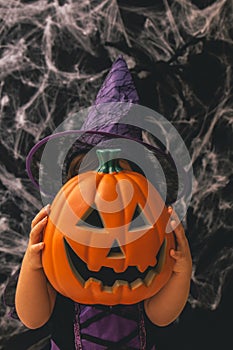 Little girl dressed as a witch holding a pumpkin against a dark background with spiderwebs