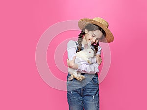 Little girl dressed in a jeans and straw hat holding a little white lovely rabbit, isolated pink background.