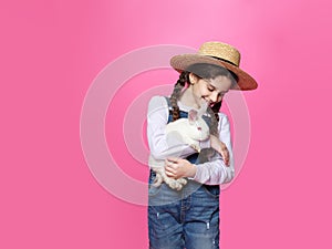 Little girl dressed in a jeans and straw hat holding a little white rabbit, isolated pink background.