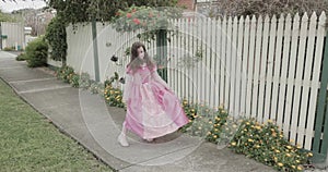 A little girl dressed in a princess costume dances, spins and twirls outside her house in front of a fence and flowers