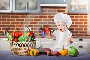 Little girl dressed in white chef hat and apron, sits among vegetables and holds an apple.