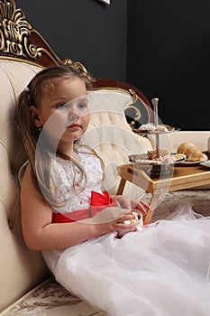 Little girl drinking tea. A child in a white dress is sitting on the royal sofa with sweets. Children& x27;s