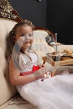 Little girl drinking tea. A child in a white dress is sitting on the royal sofa with sweets. Children& x27;s