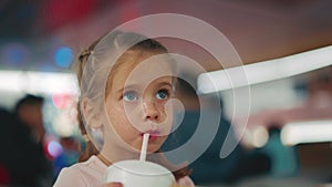 Little girl drinks milkshake from plastic glass with straw while sitting table in restaurant in the evening with light